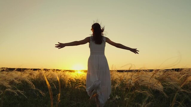 Holiday running girl. Free girl runs happily through meadow in grass in sunset. Concept of female dreams, success, travel, flight. Young woman holding her arms to sides runs across field in sun, sky