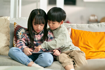 two asian children brother and sister sitting on family couch playing computer game using digital tablet
