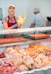 Young woman butcher in apron lays out raw chicken meat on counter in butcher shop