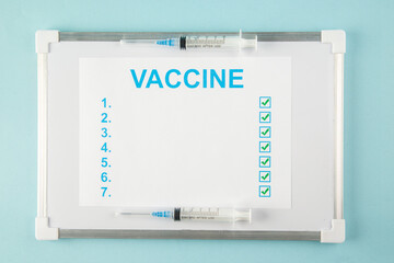Above view of a sheet with VACCINE numbers ticks and disposable syringes on white desk on blue background