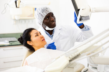 Asian woman sitting on dental chair. African-american man dentist pointing at display.