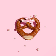 Heart shaped pretzel, creative aesthetic food concept, satin bow, pearls, festival days idea, pastel pink background.