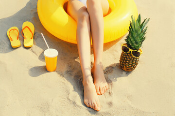 Summer vacation, close up legs of relaxing woman lying on sand on the beach with yellow swimming inflatable ring, pineapple in sunglasses, cup of juice, flip flops