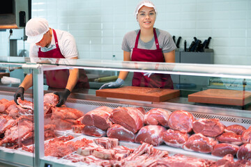 Adult man and young woman sellers in uniform display raw meat beef in butcher shop..