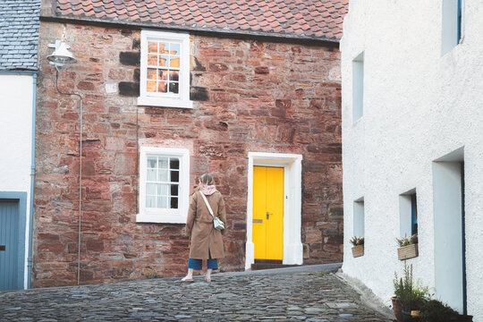 A young blonde female tourist explores a quiet cobblestone lane in the quaint and charming old town of the seaside coastal fishing village of Crail, Fife, Scotland, UK.