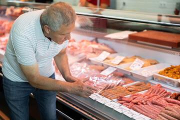 Elderly man buyer of meat department of grocery store, looking at meat small sausage in refrigerator window. Male pensioner customer points with finger and orders chorizo meat snacks