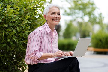Smiling senior gray haired business woman using laptop computer, typing on keyboard sitting in...