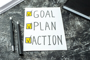 Front view of white sheet with GOAL PLAN ACTION writings ticks pens and notebook on black gray distressed background