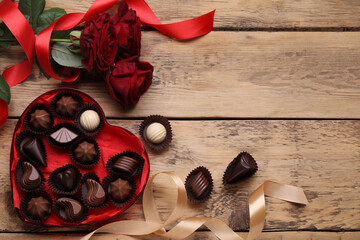 Heart shaped box with delicious chocolate candies, roses and ribbon on wooden table, flat lay....