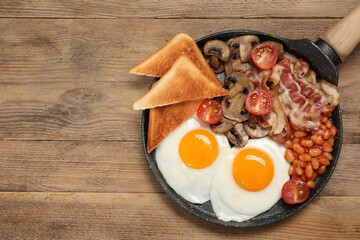 Serving pan of fried eggs, mushrooms, beans, bacon, tomatoes and toasted bread on wooden table, top view with space for text. Traditional English breakfast