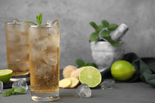 Glass of tasty ginger ale with ice cubes and ingredients on grey wooden table, space for text