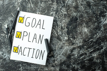 Overhead view of white blank with GOAL PLAN ACTION writings and ticks pen on notebook on black gray distressed background with free spaces