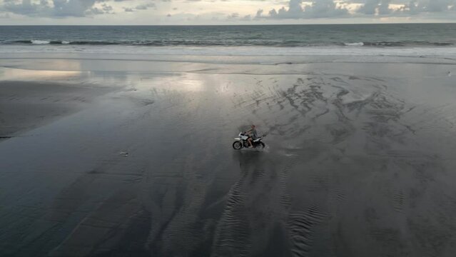 Aerial: Man rides motorcycle through shallow water on wide sand beach