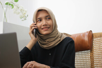 Beautiful woman in hijab in office, happy and smiling Southeast Asian business woman calling someone using her smartphone.
