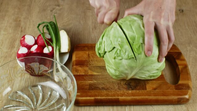 A woman uses a kitchen knife to cut fresh white cabbage on a wooden cutting board. Close-up female hands chopping cabbage for making summer salad. The concept of cooking food in the home kitchen
