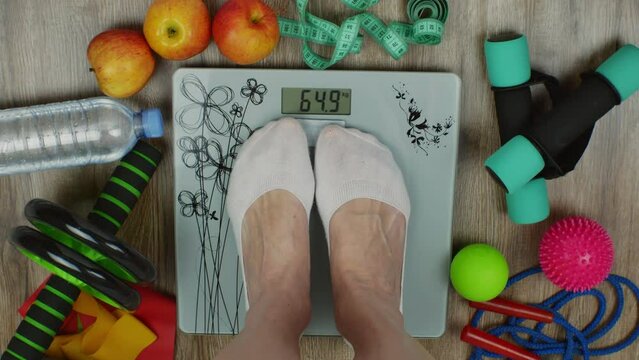 Floor scales with female feet standing, apples, water bottle, measuring tape, dumbbells, fitness rubber bands, a ball, acupuncture balls, a skipping rope lie around. Concept of overweight and obesity