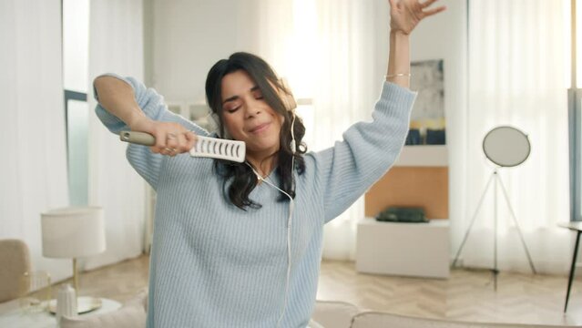 Slow motion funny hispanic woman dancing moving hands. Joyful people lifestyle concept, dancing singing in hair brush mic. Portrait of happy crazy carefree latin american girl having fun in apartment
