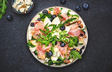 Gourmet salad with sweet pears, blueberries, blue cheese, smoked pork ham? arugula and walnuts. Black table background, top view