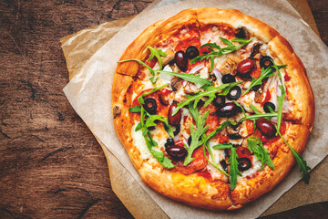 Pizza with salami sausage, mozzarella cheese, mushrooms, black olives, spicy tomato sauce and arugula, rustic wooden table background, top view