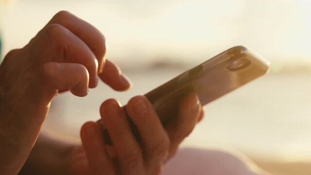 Close-up macro footage of aged female hands holding cell phone and scrolling social media against golden light on sunset at seaside. Elderly lady tapping screen of mobile phone. Leisure outdoors.