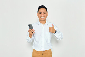 Portrait of smiling young Asian man in formal wear holding mobile phone and giving thumbs up, looking at camera with happy expression isolated on white background