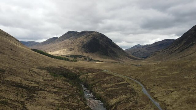 Panoramic aerial view of Scotland's rugged highlands. Remote meadow, winding road, majestic mountains, and river under a cloudy sky. Film location of Skyfall, James Bond movie.