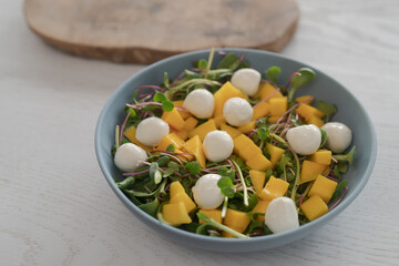 Salad with mango, mozzarella and mixed microgreens in blue bowl on white oak table