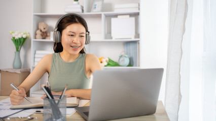 Beautiful young asian woman watching live video or video call of teacher teaching on laptop in her home, Take notes of important conversations and messages during the teacher's teaching.