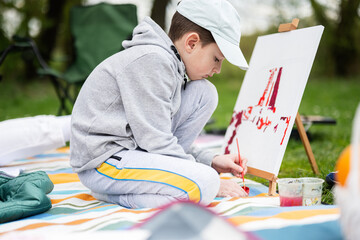 Boy enjoying outdoor on picnic blanket and painting on canvas at garden spring park, relaxation.