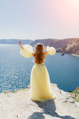 Woman yellow dress sea. Side view Young beautiful sensual woman in yellow long dress posing on a rock high above the sea at sunset. Girl in nature against the blue sky
