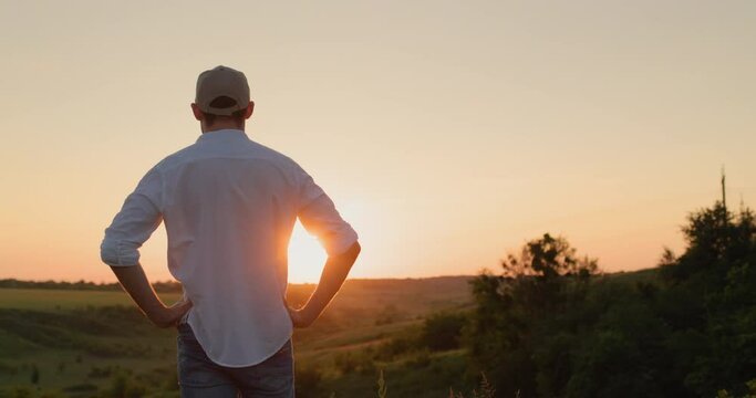 A confident young farmer stands in front of a picturesque countryside as the sun sets. View from the back