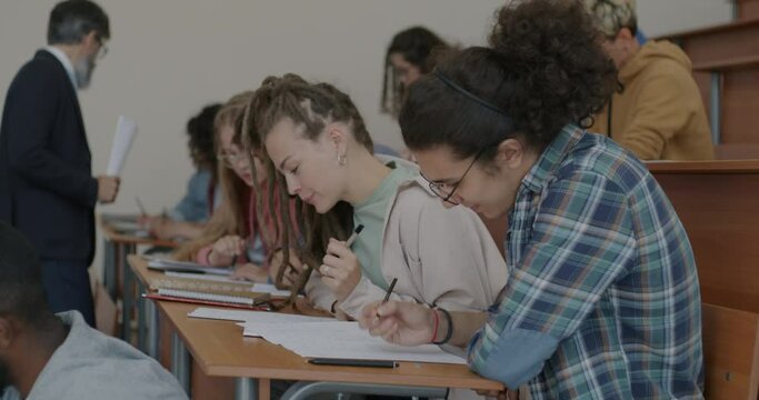 Group of young people writing test studying sitting at desks in classroom while teacher walking. College education and knowledge assessment concept.