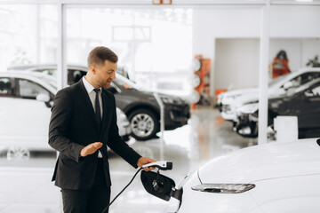 Portrait of a confident sales manager in the suit near the car charging station, selling electric...