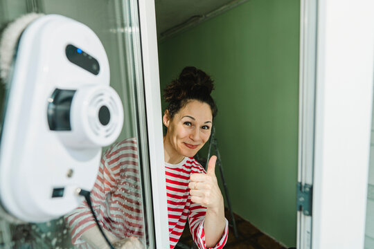 Happy woman showing thumbs up gesture and cleaning window with robot washer
