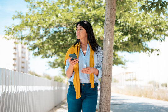 Thoughtful businesswoman holding smart phone leaning on tree