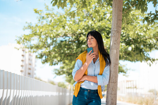 Thoughtful businesswoman leaning on tree holding smart phone