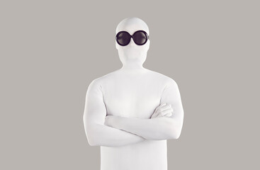 Portrait of person in bodysuit disguise and sunglasses. Man with secret, hidden personality, wearing white spandex costume and black round glasses standing with folded arms on grey color background - obrazy, fototapety, plakaty