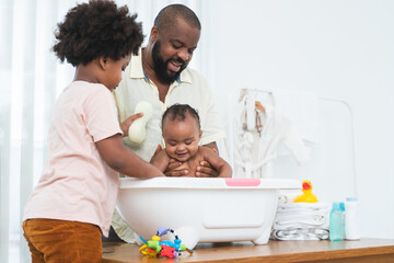 Obraz na płótnie Canvas African middle aged father bathing adorable newborn baby daughter in bathtub at home. Child boy help dad cleaning, washing little sister with sponge in bath. Kid hygiene and health care with family