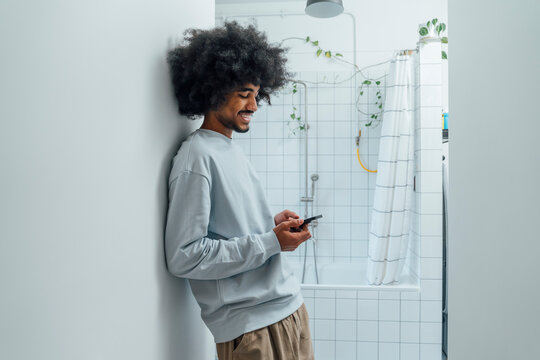 Smiling man using smart phone leaning on wall in bathroom at home
