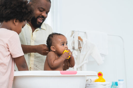 African middle aged father bathing adorable newborn baby daughter in bathtub at home. Son help dad cleaning little sister. Selective focus on little girl biting duck toy. Child hygiene and health care