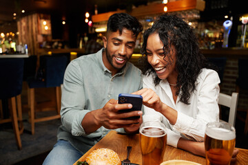 Young multiracial couple in casual clothing smiling while typing on mobile phone at restaurant