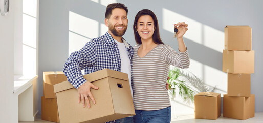 Happy young couple holding a key and cardboard boxes. Man and woman standing in new apartment looking at camera and smiling celebrating moving day. Relocating, real estate, mortgage concept. Banner.