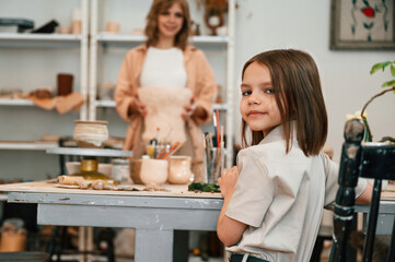 Little girl is sitting by the table in the workshop, mother holding big unfinished pot