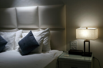 Interior of stylish bedroom with white blankets on bed and glowing lamp at night. Blue pillows on white bed in natural bedroom interior with brown lamp and wooden modern bedside table, bed. Night time