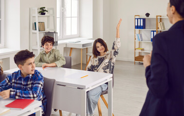 Smiling teenager schoolgirl sitting at the desk in the classroom raising her hand up ready to...