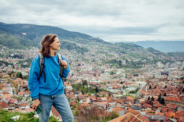 Fototapeta na wymiar A smiling traveler girl looks at a beautiful European city in the mountains from a view point. Travel to Sarajevo, Bosnia and Herzegovina