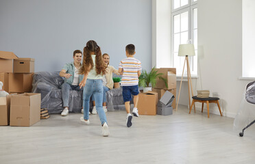 Fototapeta na wymiar Happy family enjoying moving day and having fun in their new home. Cheerful mom, dad and children running and playing games in large living room full of cardboard boxes in new big house or apartment