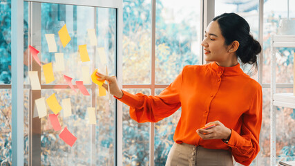 Business people work on project planning board in office and having conversation with coworker friend to analyze project development . They use sticky notes posted on glass to make it organized. Jivy