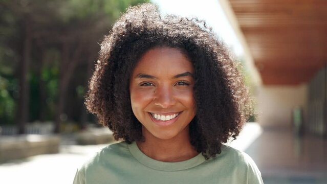 Smiling happy cute cheerful pretty young positive beautiful African American ethnic woman model with curly afro hair looking at camera in city street outdoors on sunny day, close up face portrait.