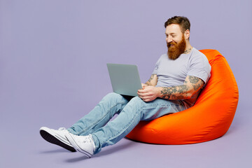 Full body young redhead bearded man wear violet t-shirt casual clothes sit in bag chair hold use work on laptop pc computer isolated on plain pastel light purple background studio Lifestyle concept